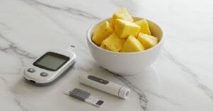 A small white bowl of fresh pineapple chunks sits on a clean counter next to a glucose meter, illustrating how diabetics can safely eat pineapple